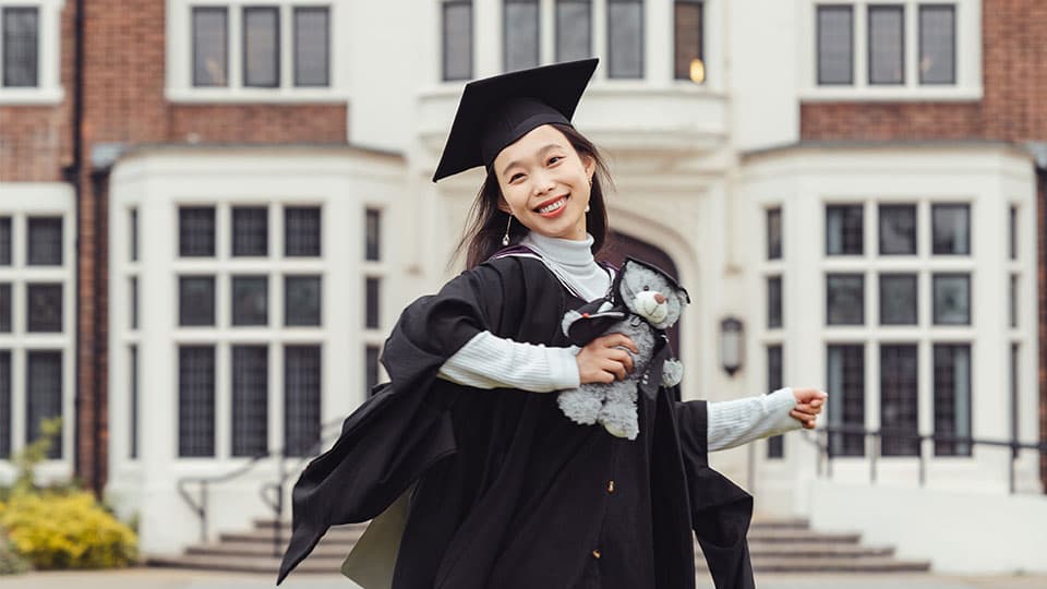 Xu wearing a graduation gown and cap holding her degree scroll outside Hazlerigg Building on 麻豆視頻_麻豆直播_麻豆传媒官网 campus. She is smiling and she is also holding a grey stuffed teddy bear which is also wearing a black cap and gown.
